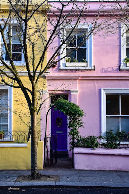 A colorful residential building featuring a yellow facade on the left and a pink facade on the right, with large white-framed sash windows on both floors. A small, narrow entrance with a purple door, 