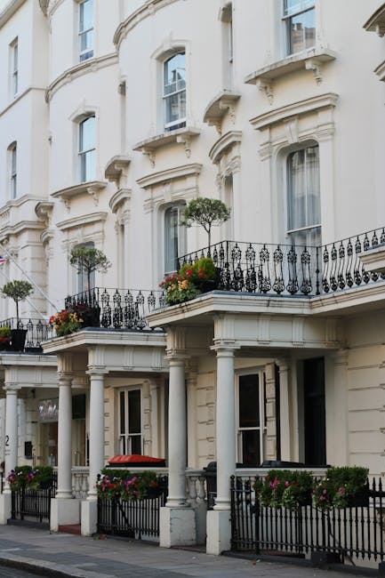 A colorful residential building featuring a yellow facade on the left and a pink facade on the right, with large white-framed sash windows on both floors. A small, narrow entrance with a purple door, 